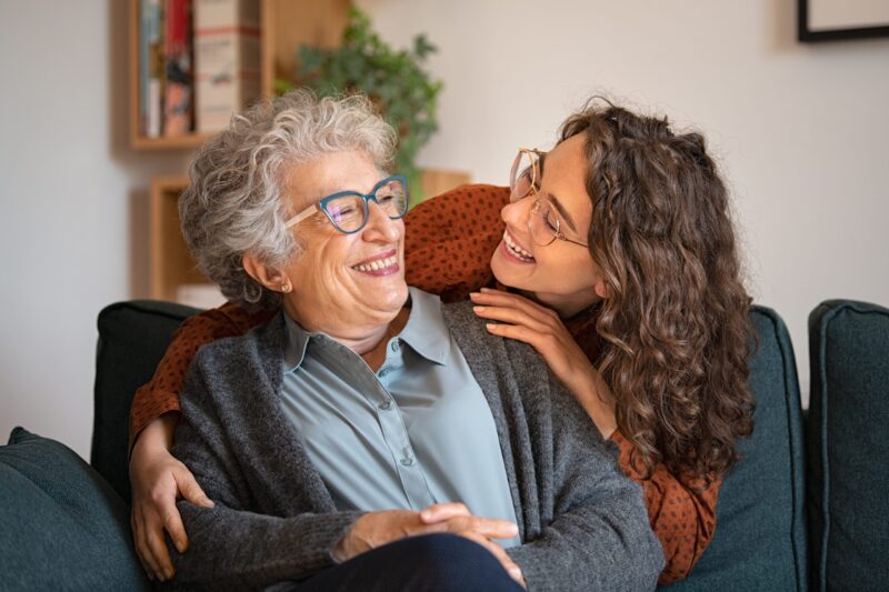 Smiling, older woman sitting on sofa being embraced by smiling, younger woman