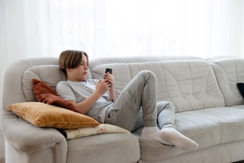 Teenaged boy lounges on a gray sofa while looking at his cell phone.