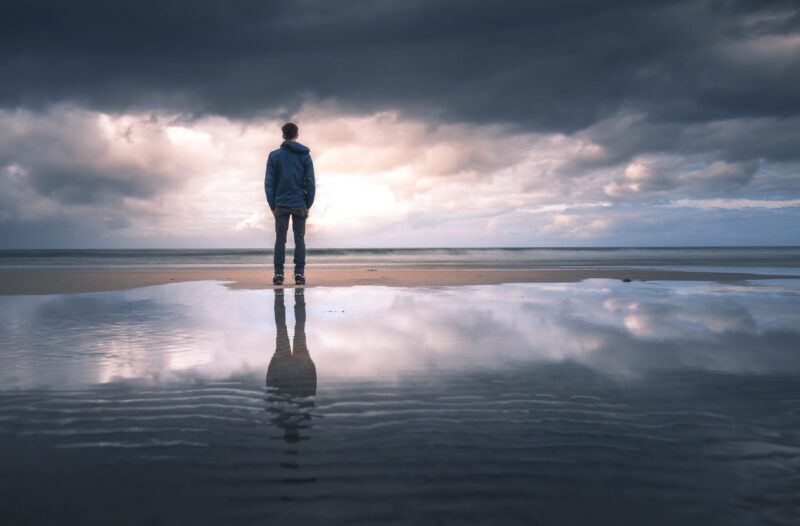A man stands at the shoreline, his back to the camera, looking out over the horizon with gray clouds descending at sunset.