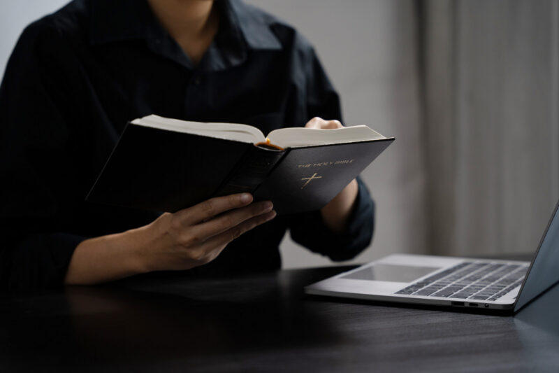 Man holding open Bible with laptop on table