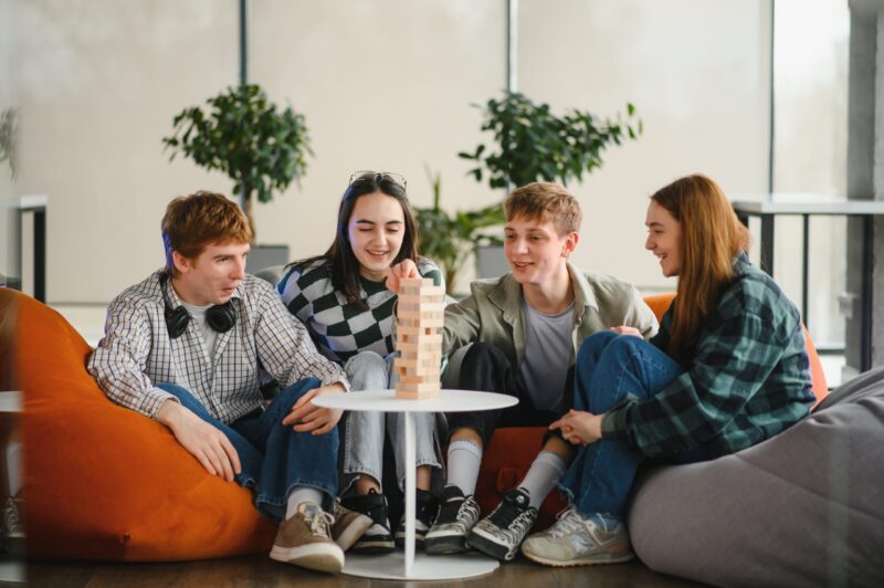 Four cheerful friends are sitting on beanbag chairs, enjoying a friendly board game, building a tower together, and sharing laughter