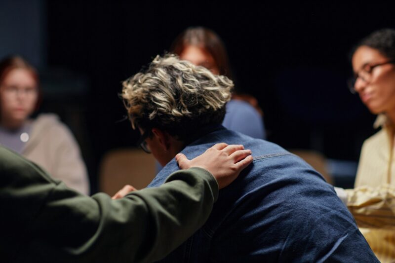 Young man with face down and a hand on his shoulder in a circle of people praying for him